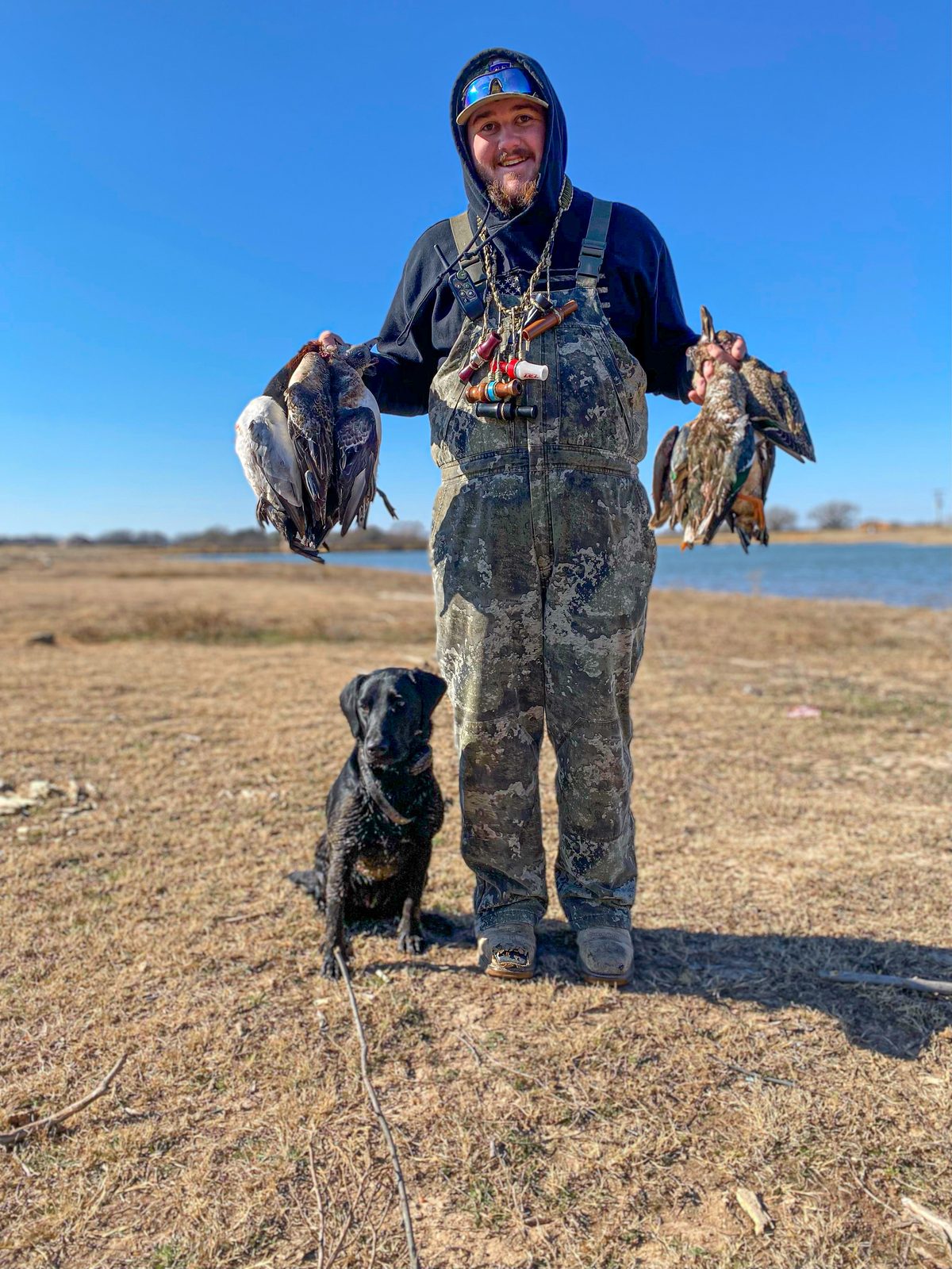 Guide Hunter Murphy holding a strap of ducks in each hand beside a pond, with his black lab sitting at his feet